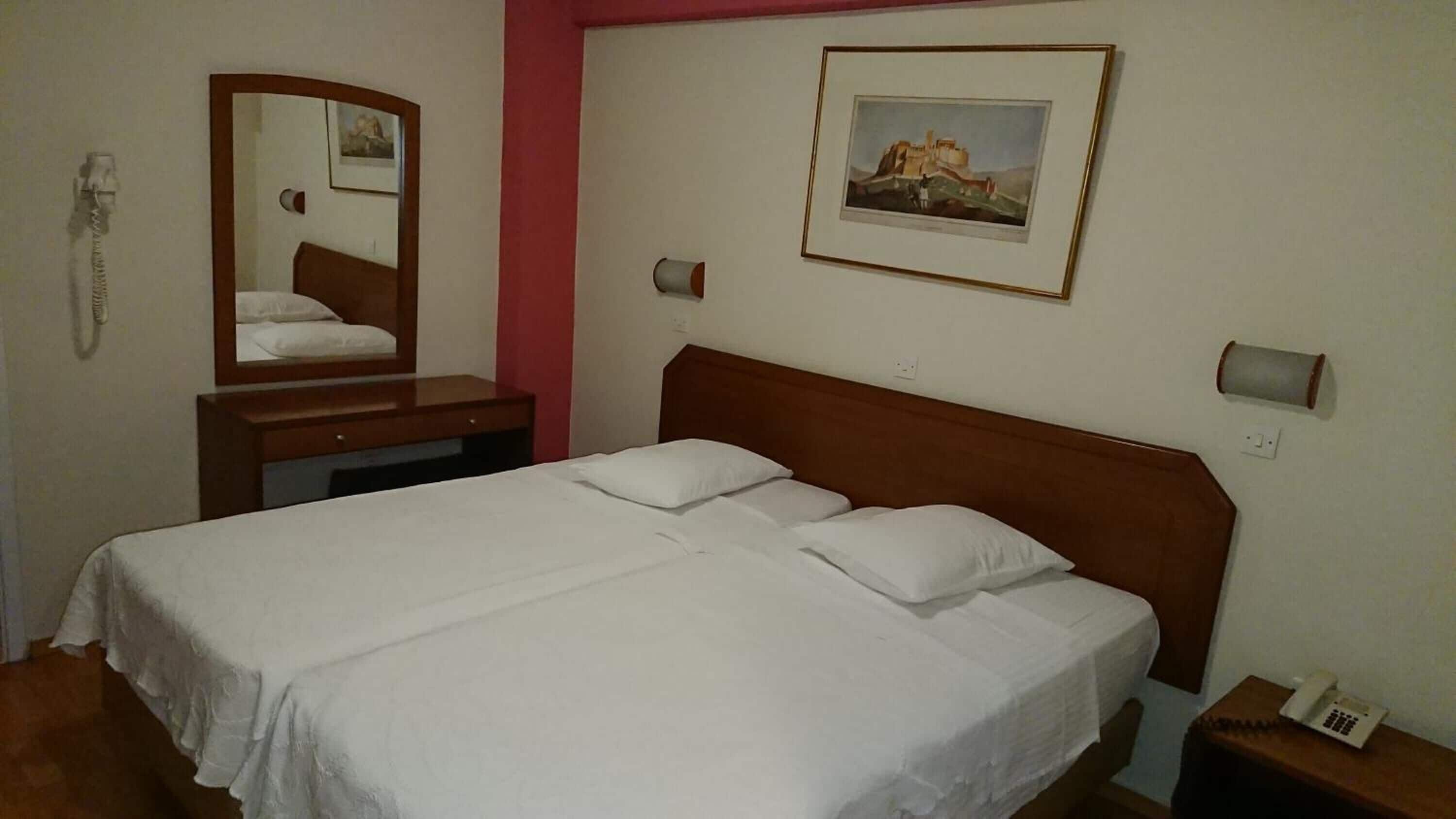 A hotel room with a bed, a bedside table, a mirror, a wall-mounted phone, and a framed picture.