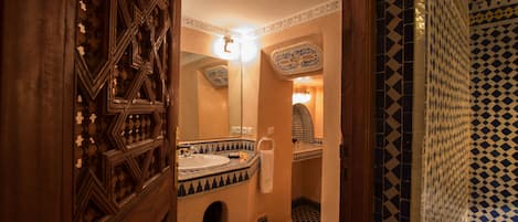 A traditional Moroccan bathroom with intricate tile work, a wooden door, and a small alcove with a mirror and light fixture.