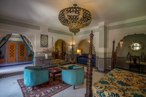 A traditional Moroccan room with a large bed, two blue armchairs, a wooden floor, and intricate tile work.