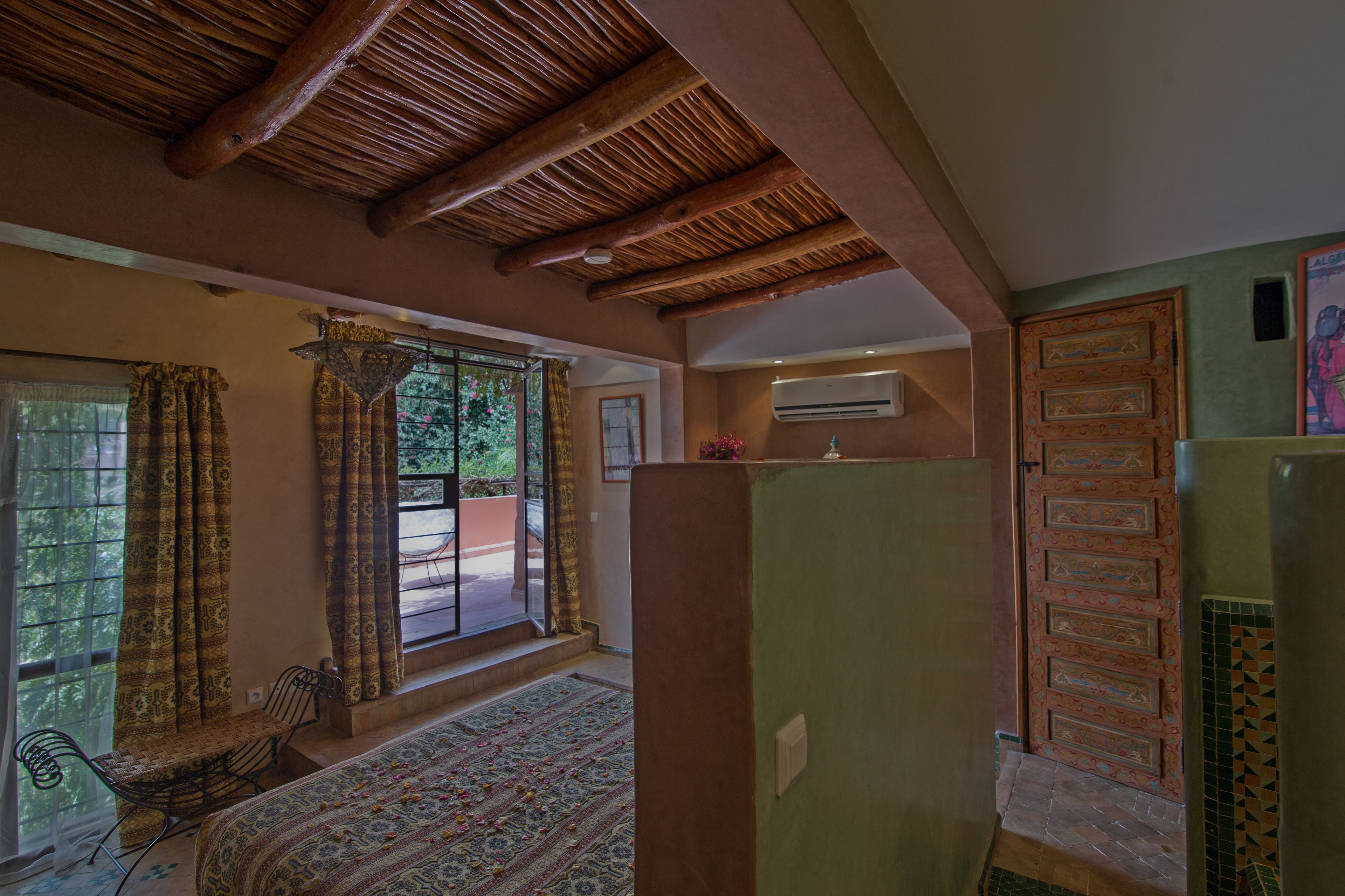 A room with a bed, a chair, a window with curtains, a door, and a ceiling with wooden beams.