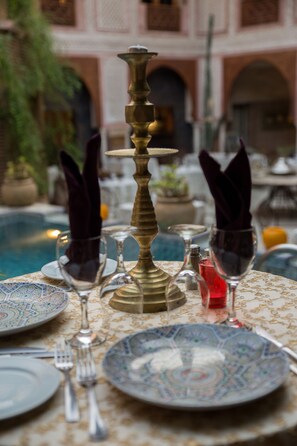 A dining table set with plates, glasses, and cutlery, surrounded by a pool and a building with arches.