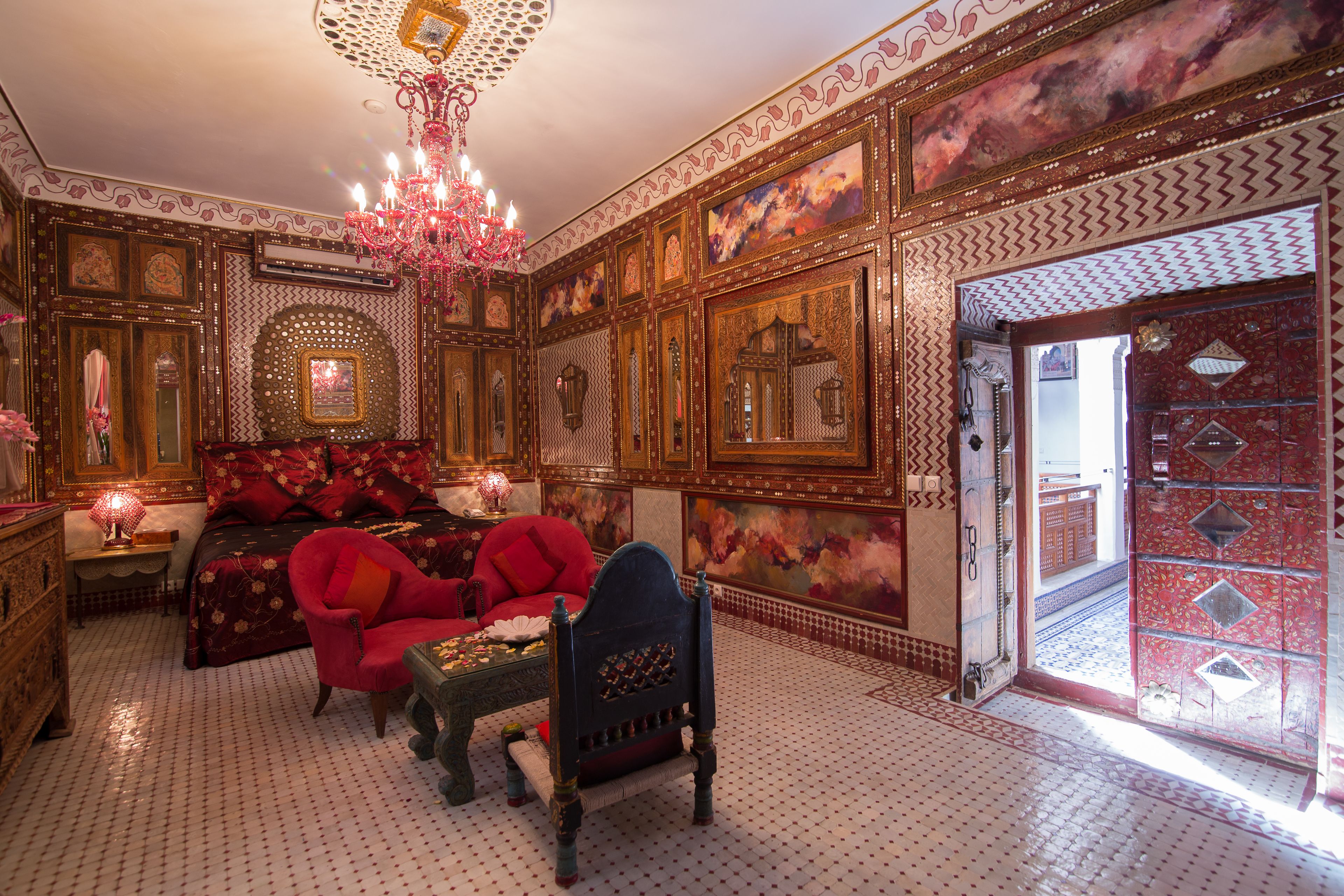 Una stanza riccamente decorata con pareti elaborate, un lampadario, un letto con biancheria rossa e una poltrona rossa.