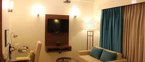 A living room with a flat-screen TV, a sofa with blue cushions, a glass coffee table, a lamp, and a wall-mounted shelf.