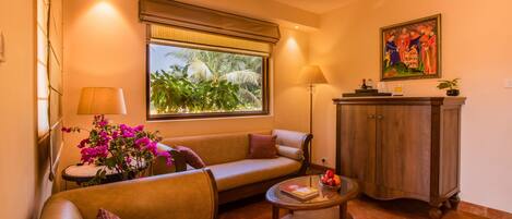 Un salón con sofá, sillón y una pequeña mesa con un libro y fruta. Hay una ventana con vistas a la vegetación y un cuadro en la pared.