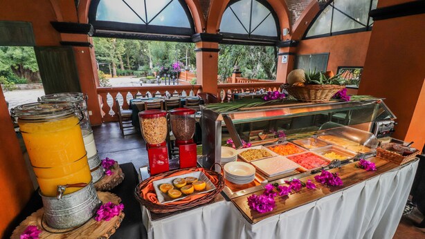 Ein Buffet mit verschiedenen Speisen und Getränken, darunter ein großer Orangensaftautomat, Schüsseln mit Müsli und ein Glas Milch.
