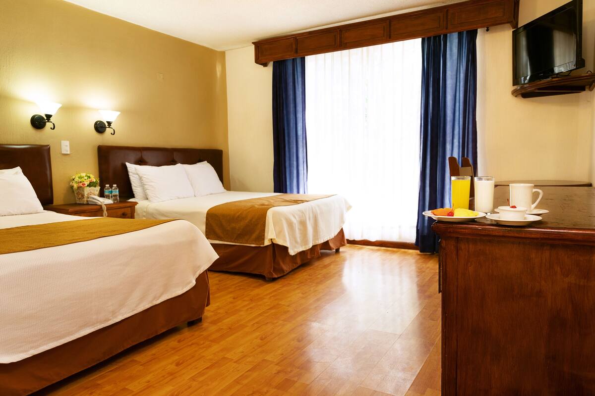 Ein Hotelzimmer mit zwei Betten, Holzboden, einem Schreibtisch mit Frühstücksangeboten und einem an der Wand montierten Fernseher.