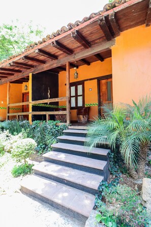 Un edificio con un porche de madera, escalones que conducen a una puerta y un jardín con plantas y una palmera.