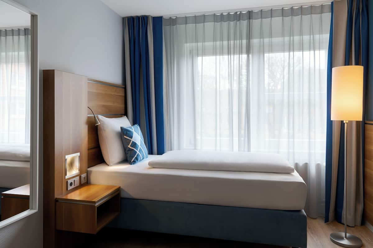 Habitación de hotel con cama, mesita de noche, lámpara y un amplio ventanal con cortinas.