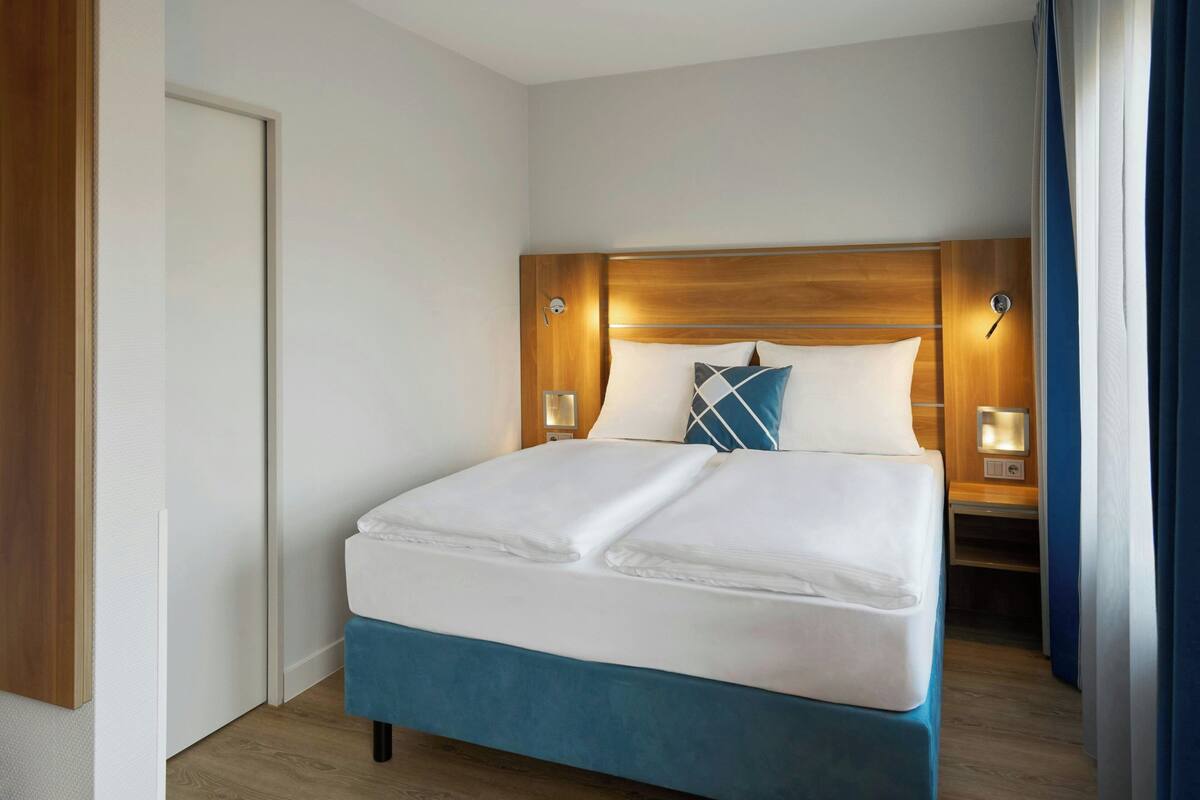 Una habitación de hotel con una cama grande, cabecera de madera, dos mesitas de noche con lámparas y una puerta.