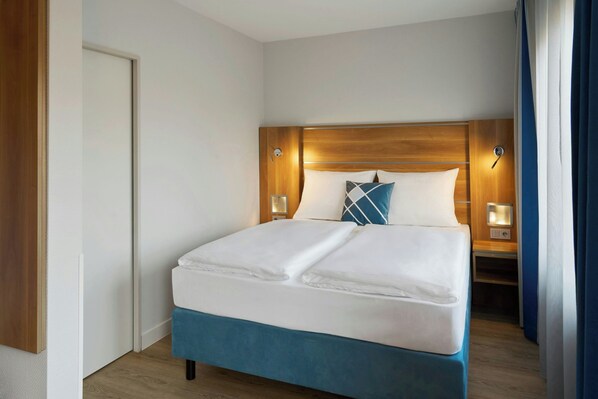 Una habitación de hotel con una cama grande, cabecera de madera, dos mesitas de noche con lámparas y una puerta.