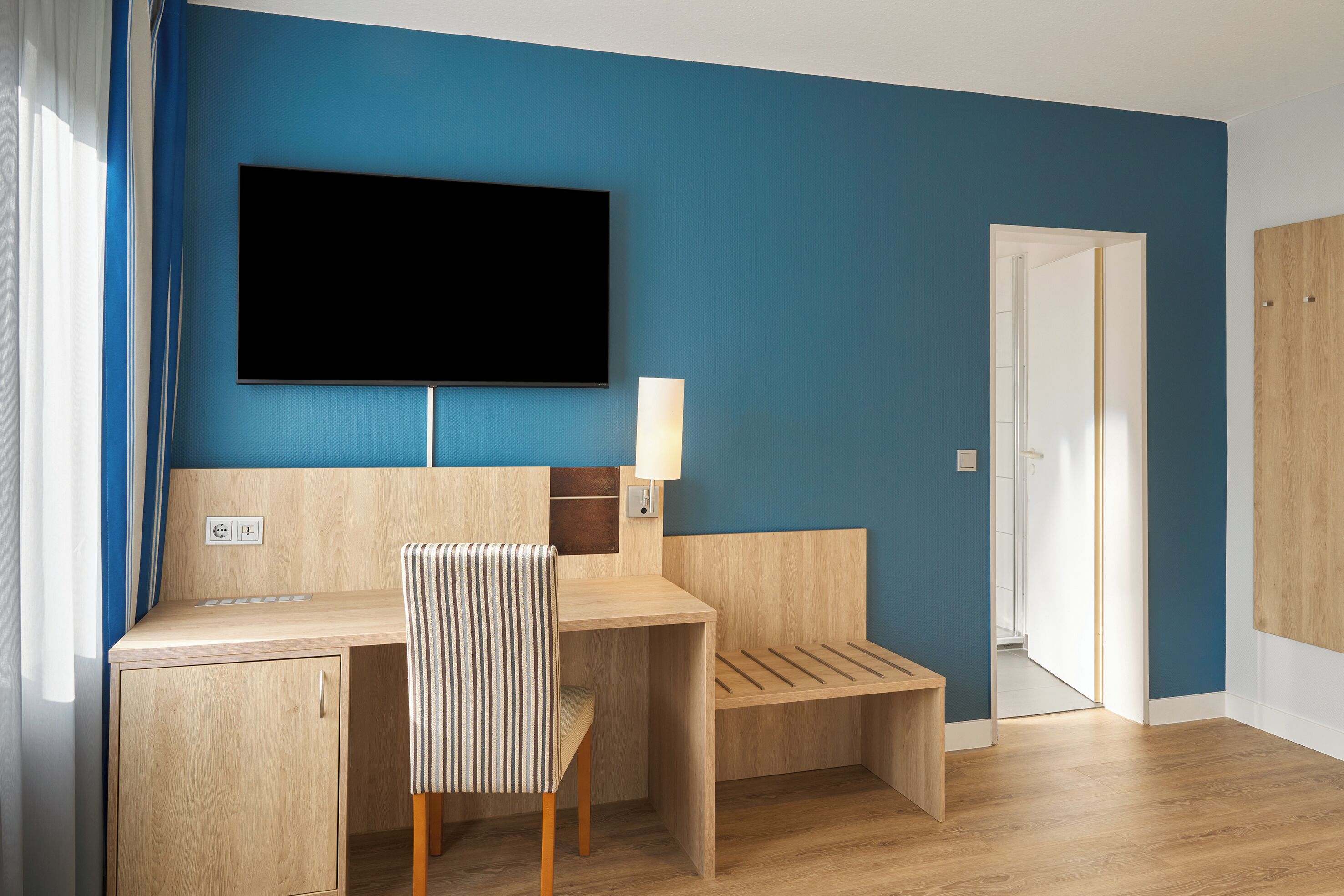 A room with a wooden desk, a chair, a TV mounted on the wall, and a door.
