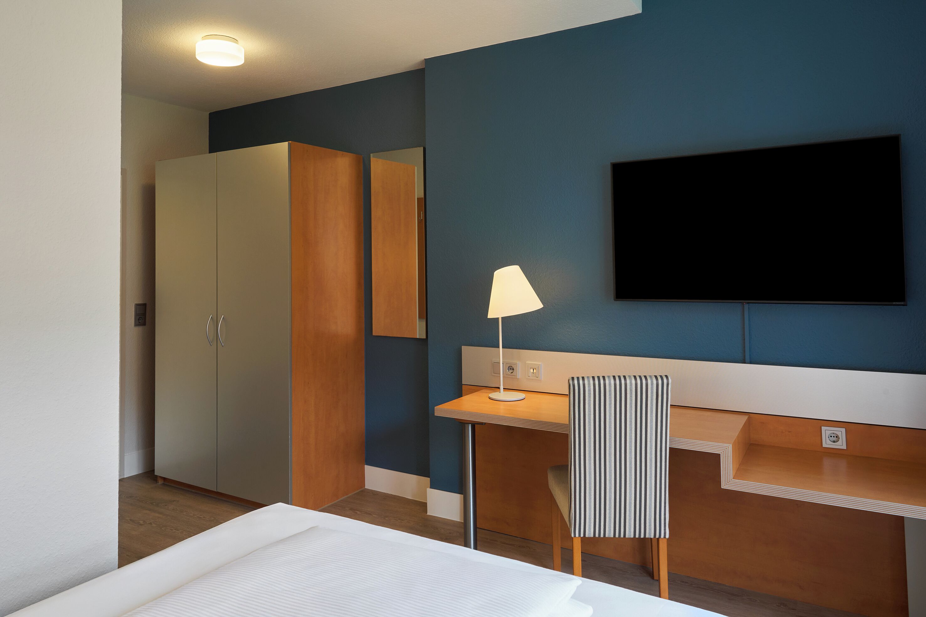 A hotel room with a bed, a desk, a chair, a TV, and a wardrobe.