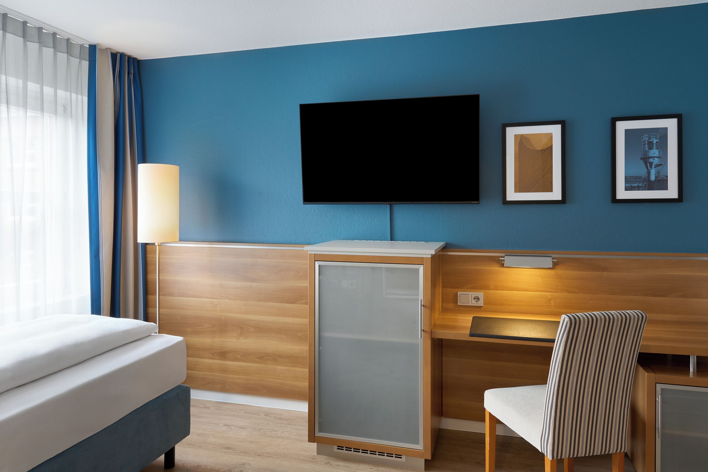 A hotel room with a bed, a desk, a chair, a TV, and framed pictures on the wall.