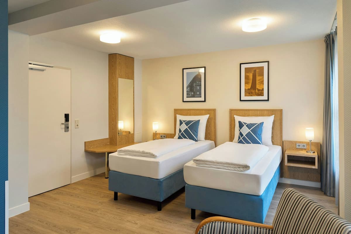 Habitación de hotel con dos camas, un banco de madera, una mesita y un estante fijado a la pared.