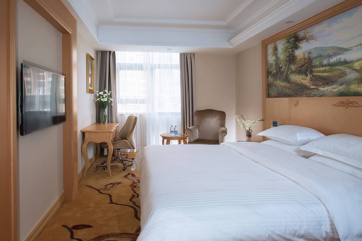 A hotel room with a large bed, a desk with a chair, a television, and a painting on the wall.