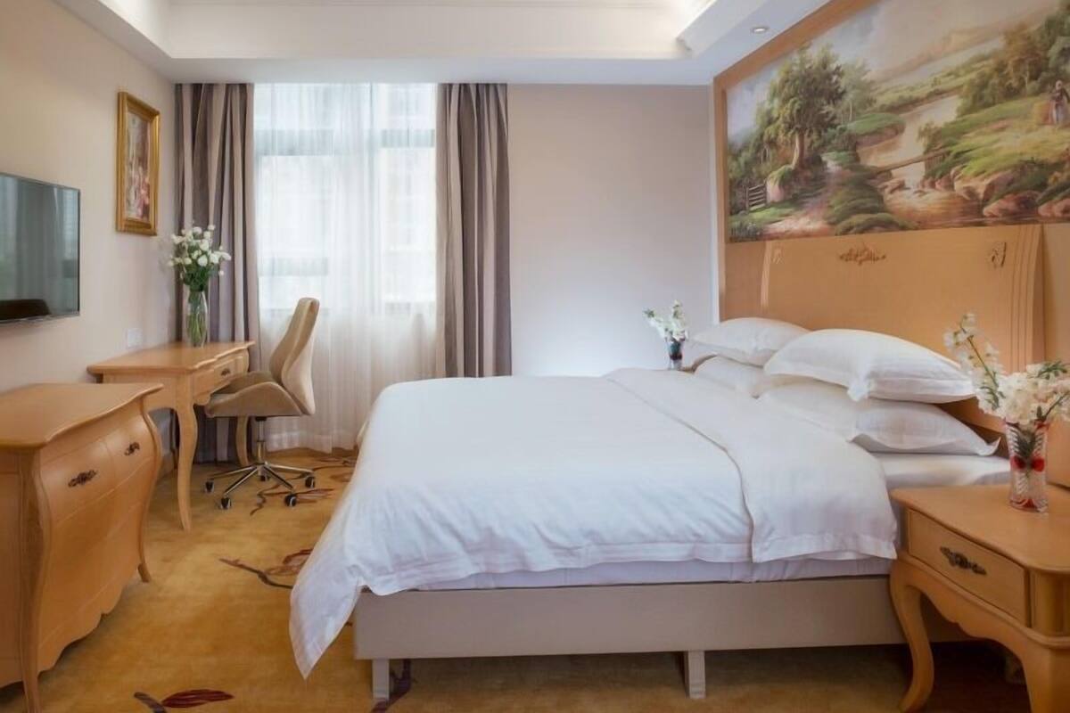 A hotel room with a bed, a desk, a chair, and a painting on the wall.