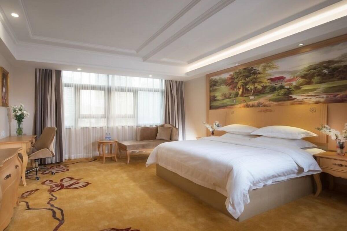 A hotel room with a large bed, a desk, a chair, and a painting on the wall.