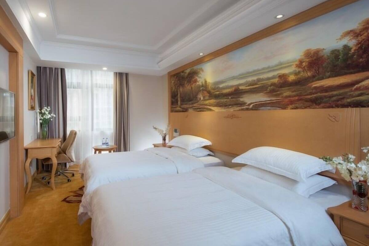 A hotel room with two beds, a desk, and a painting of a landscape on the wall.