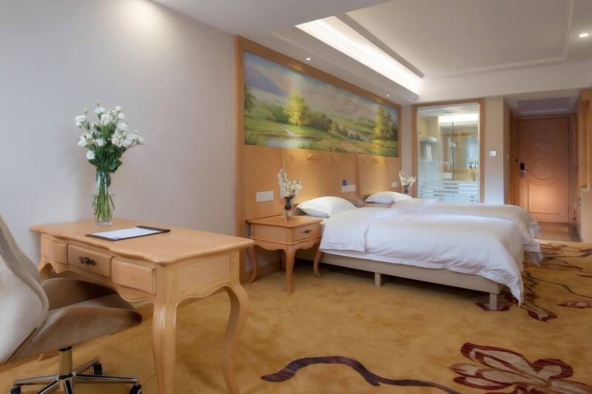 A hotel room with a bed, a desk, a chair, a vase with flowers, and a painting on the wall.