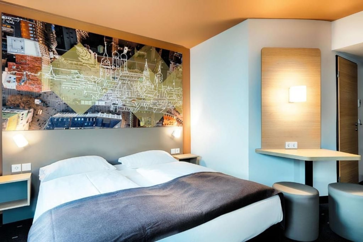 Una habitación de hotel moderna con una cama grande, un mapa colgado en la pared, una mesita pequeña y dos taburetes.