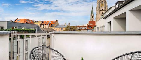 Une terrasse sur le toit, avec deux chaises en fil de fer noir et une petite table sur laquelle est posée une plante en pot. On aperçoit le clocher d’une église en arrière-plan.