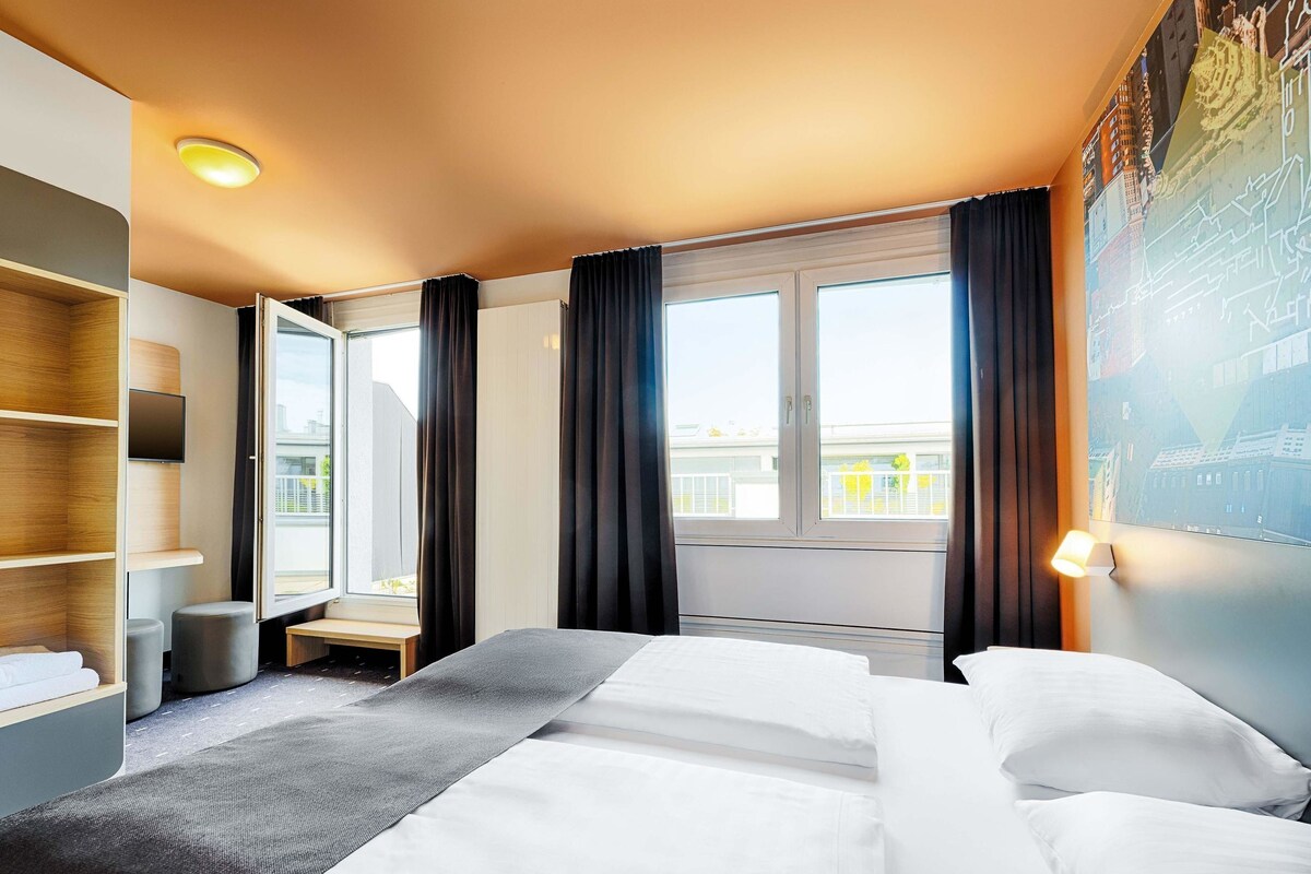 A modern hotel room with a large bed, a flat-screen TV, a small seating area, and a window with curtains.