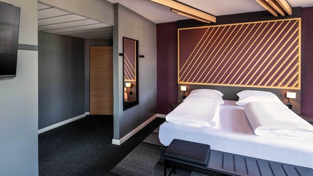 A modern hotel room with a large bed, bedside tables, a flat-screen TV, and a unique geometric wall design. - B&B Hotel München-Hbf (Munich)