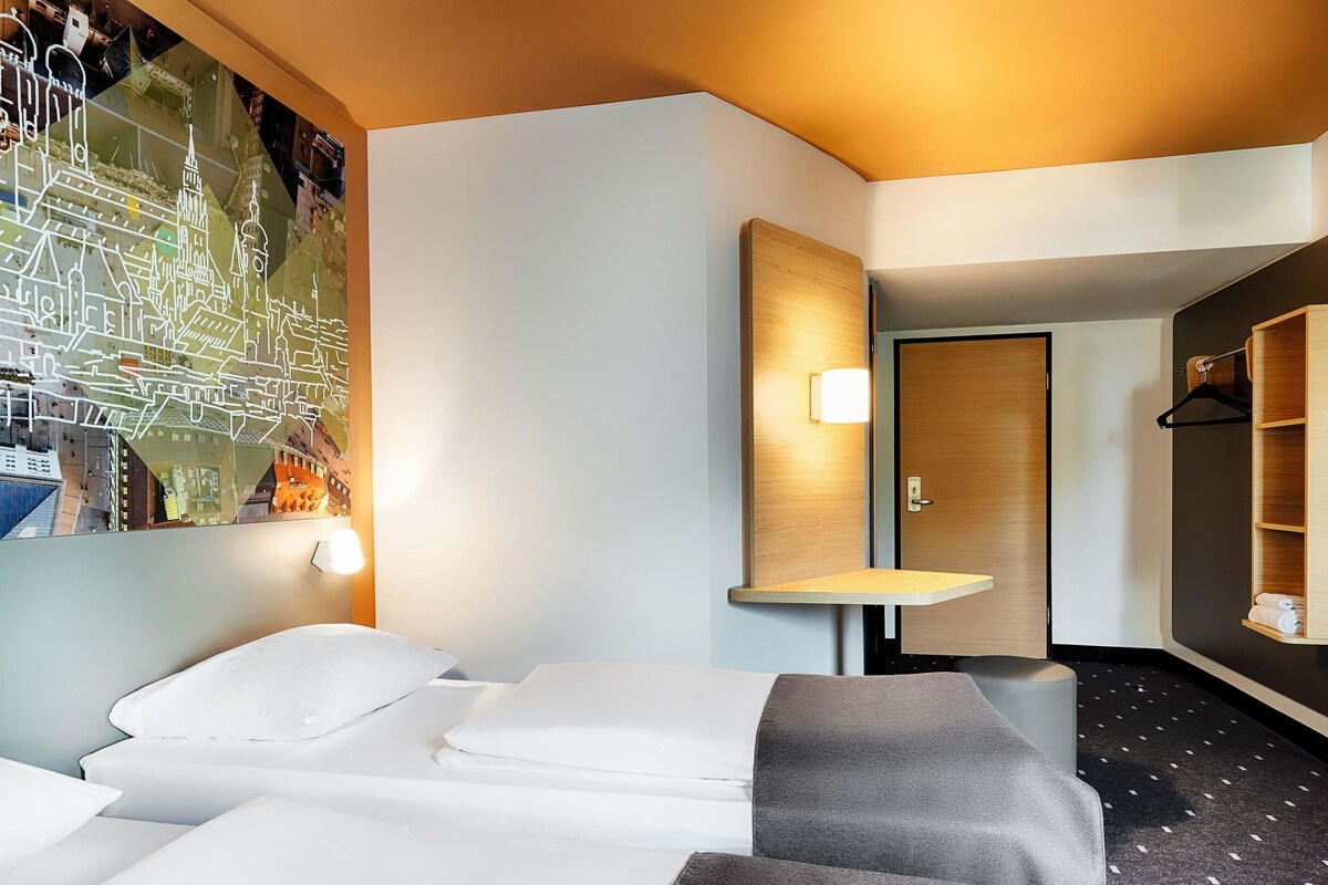 A hotel room with a bed, a desk, a mirror, and a wall with a cityscape mural.