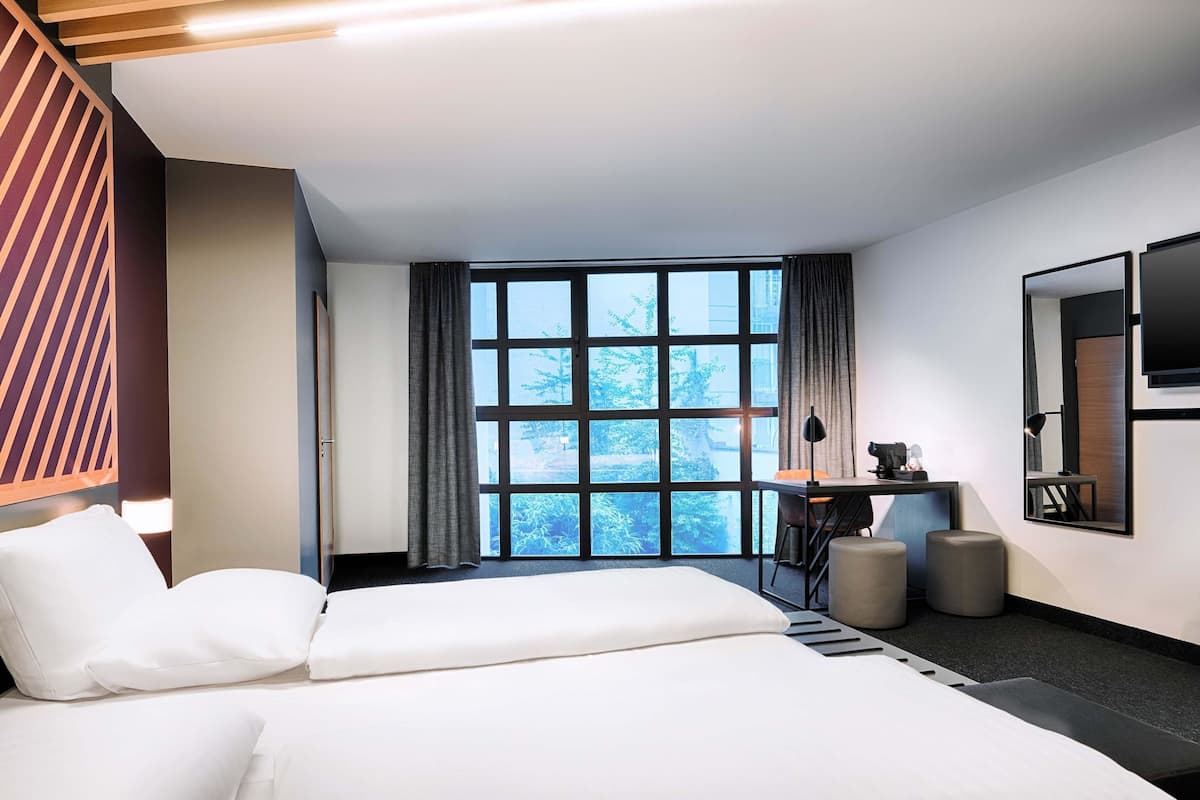 Ein modernes Hotelzimmer mit zwei Betten, einem Schreibtisch, einem Sessel, einem Fernseher und einem großen Fenster mit Blick auf Bäume.