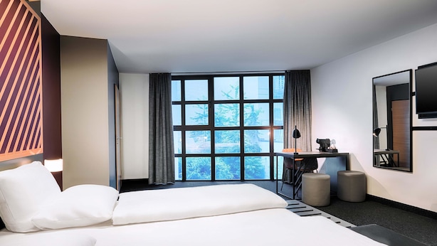 A modern hotel room with two beds, a desk, a chair, a TV, and a large window with a view of trees. - B&B Hotel München-Hbf (Munich)