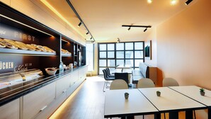 A modern cafe with a counter displaying baked goods, a seating area with tables and chairs, and large windows providing natural light. - B&B Hotel München-Hbf (Munich)