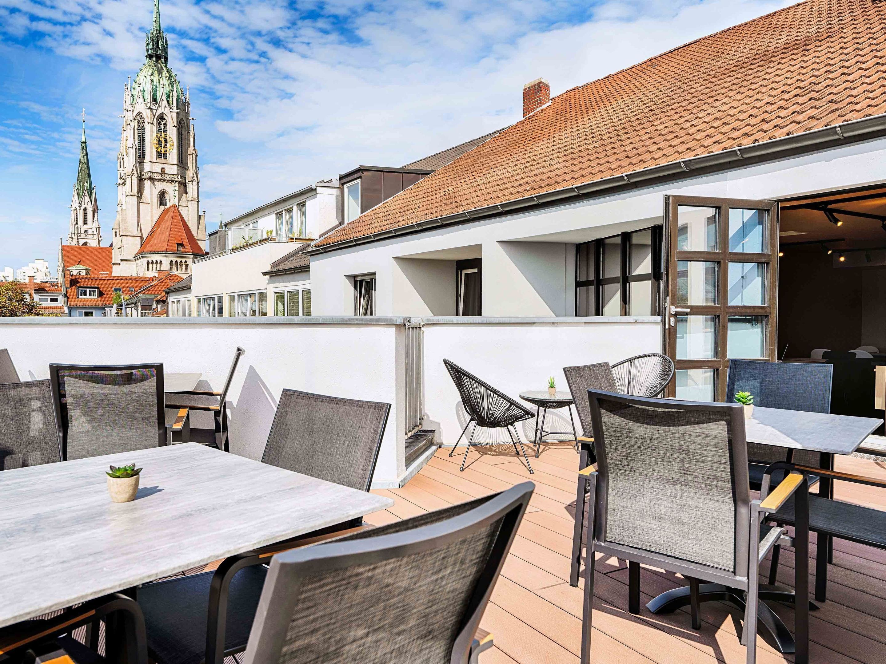 An outdoor patio with tables and chairs, a view of a church with a clock tower, and a building with large windows.