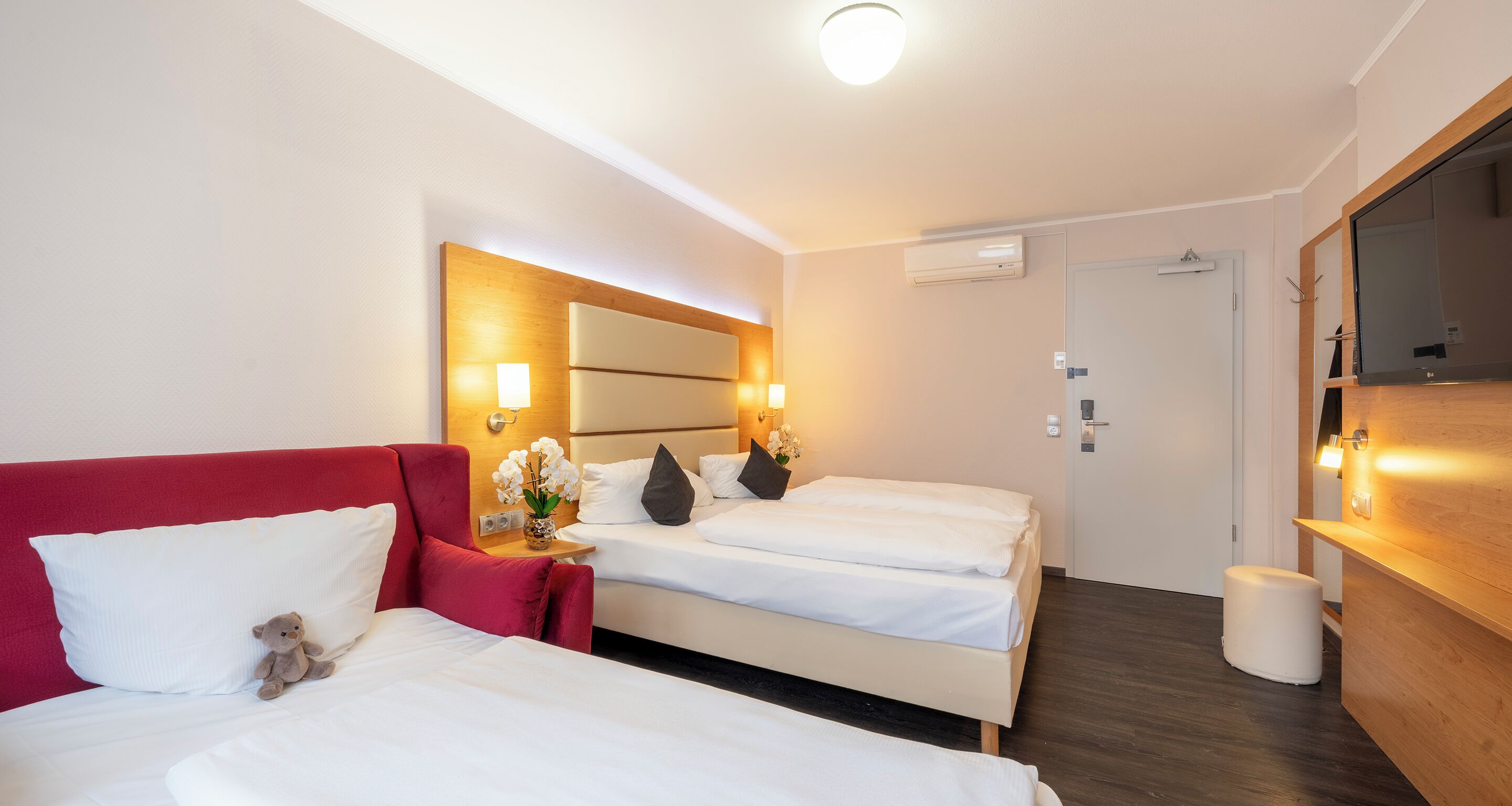 Een hotelkamer met twee bedden, een rode bank, een klein tafeltje met een bloevas en een houten kast.