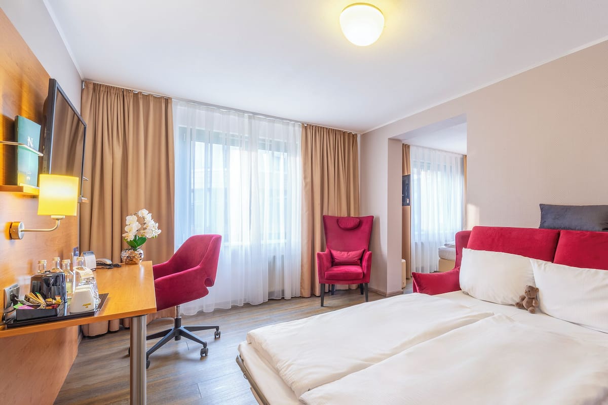 Una habitación de hotel con cama, escritorio con lámpara, una silla y un sillón rojo.
