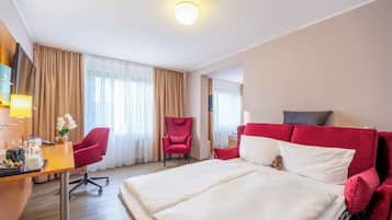Una habitación de hotel con cama, escritorio con lámpara, una silla y un sillón rojo.