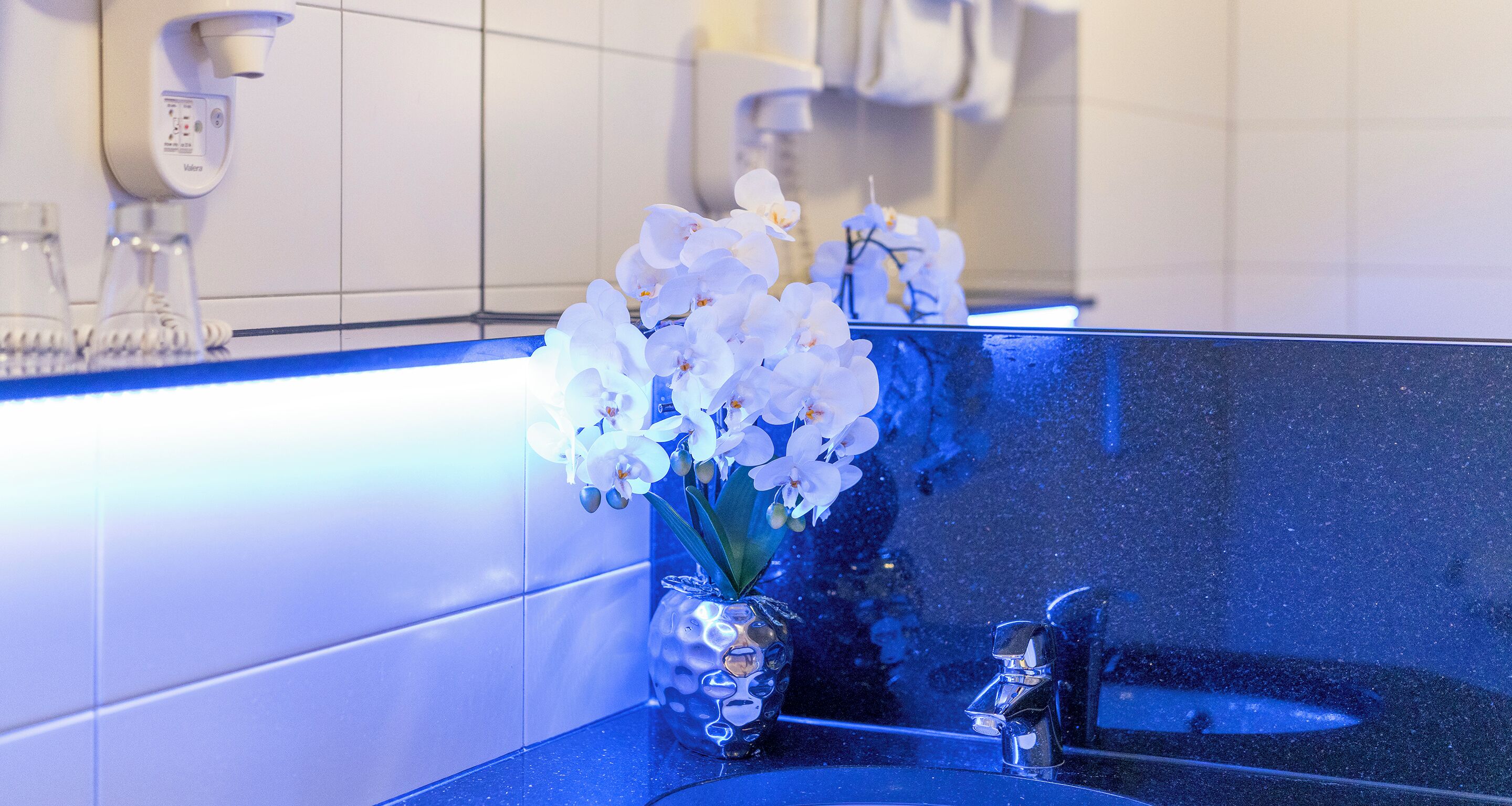 A bathroom with a blue countertop, a white orchid vase, and a mirror reflecting the bathroom fixtures.