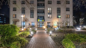 L’entrée d’un hôtel Plaza Inn la nuit, avec un chemin pavé menant aux portes, entourée de verdure et éclairée par des lampadaires.
