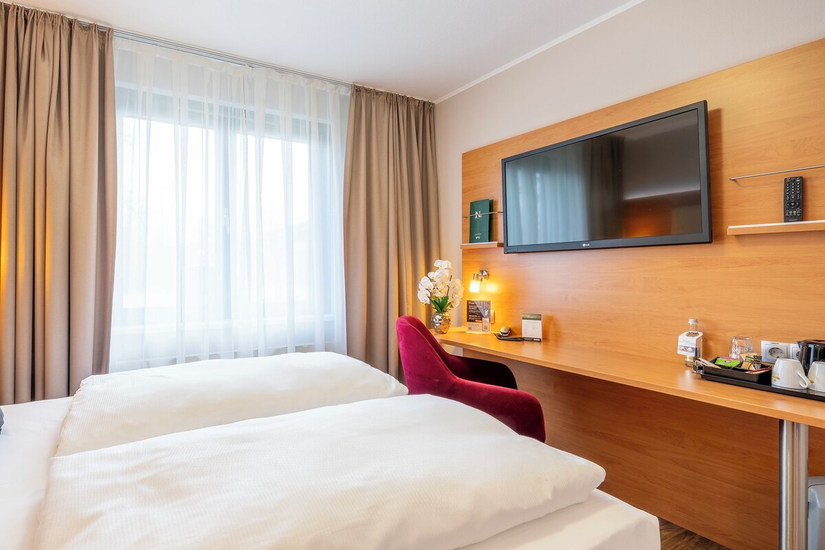 Ein Hotelzimmer mit zwei Betten, einem Fernseher, einem Schreibtisch mit Fernbedienung und einem Fenster mit Vorhängen.