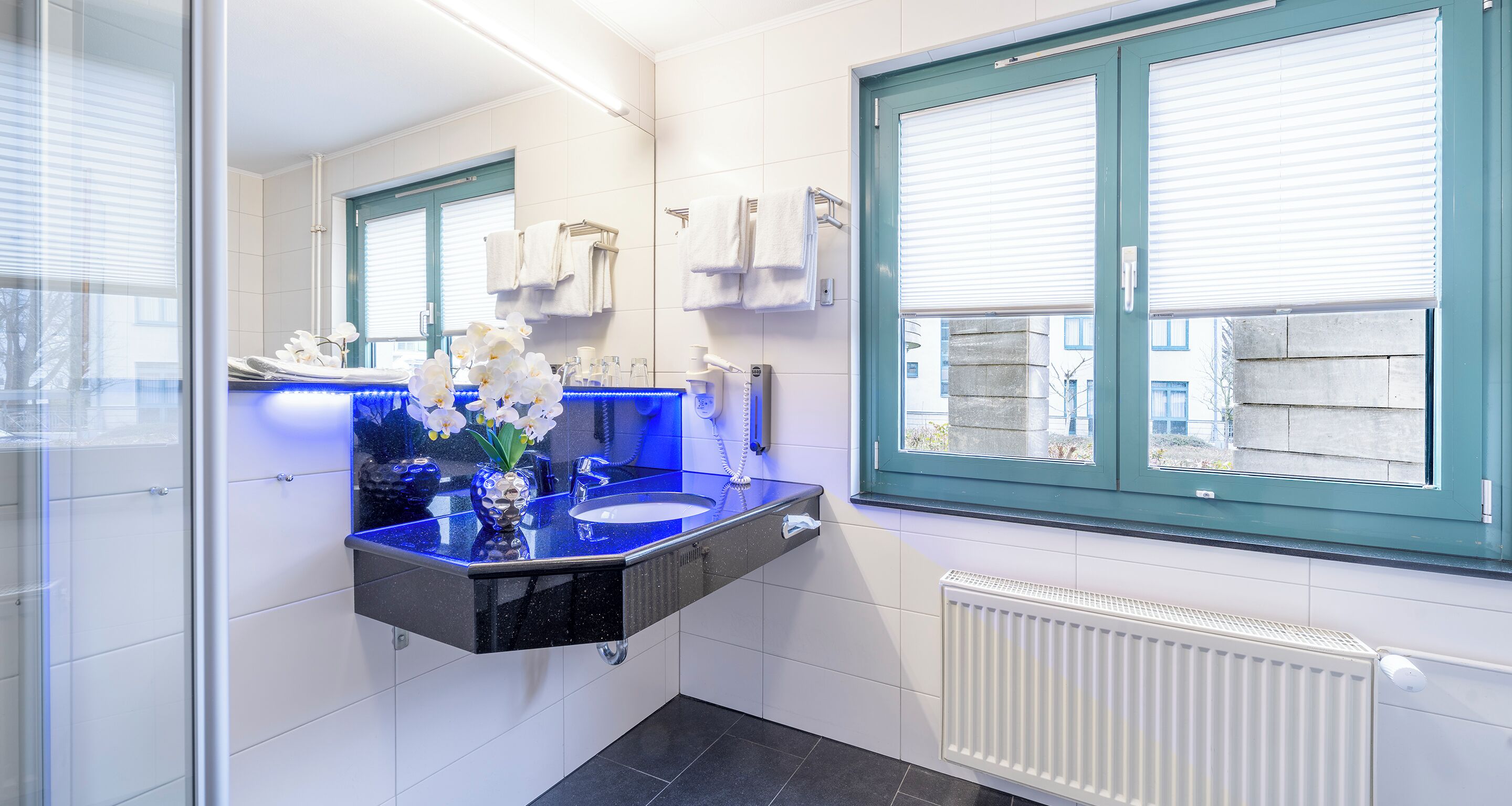 Une salle de bain moderne avec un lavabo bleu, des carreaux blancs, un grand miroir et une fenêtre avec des stores.