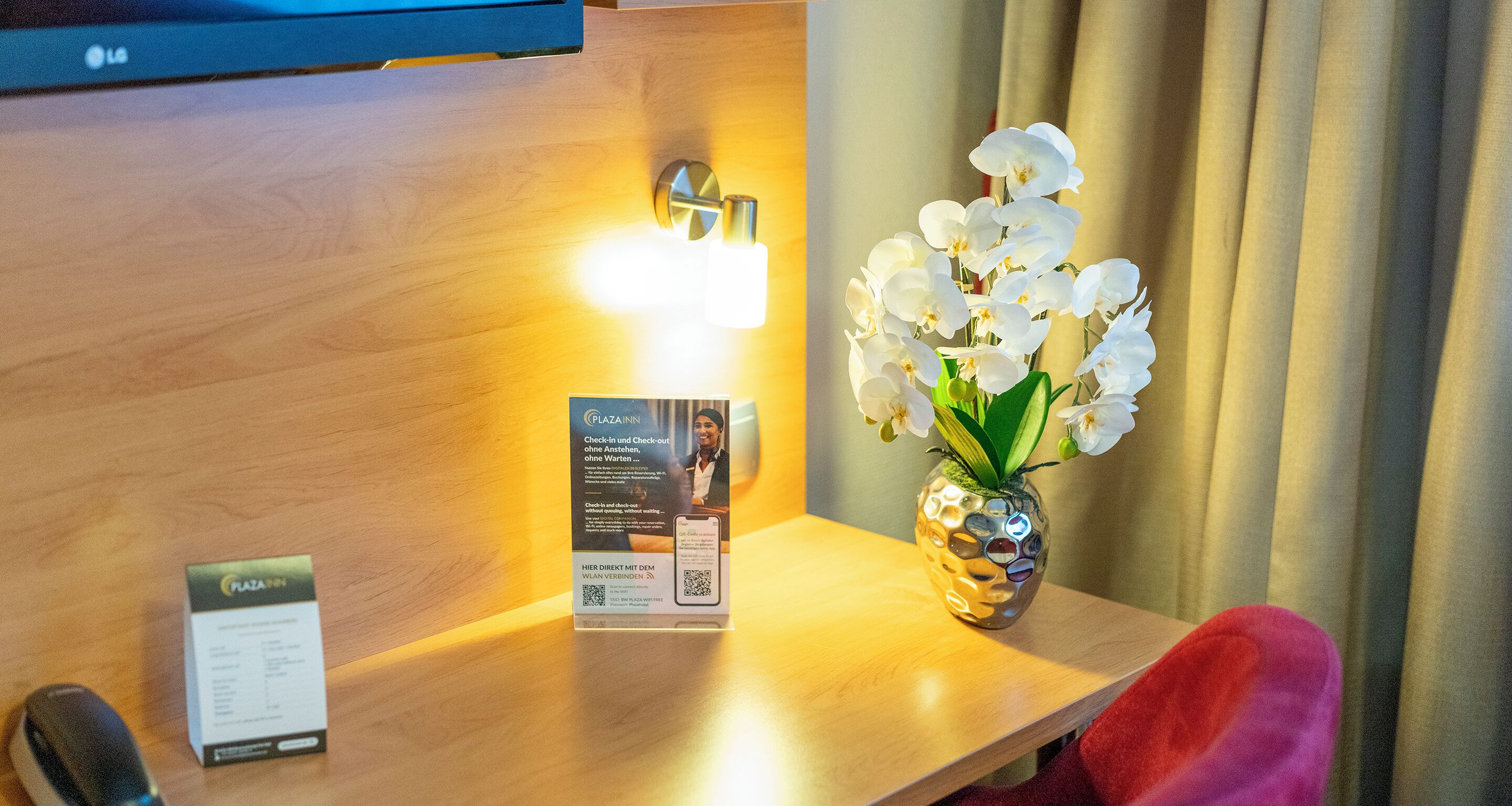 Une chambre d’hôtel avec un bureau en bois, un vase d’orchidées blanches, un présentoir pour les clés d’enregistrement et de départ, et un téléviseur à écran plat fixé au mur.
