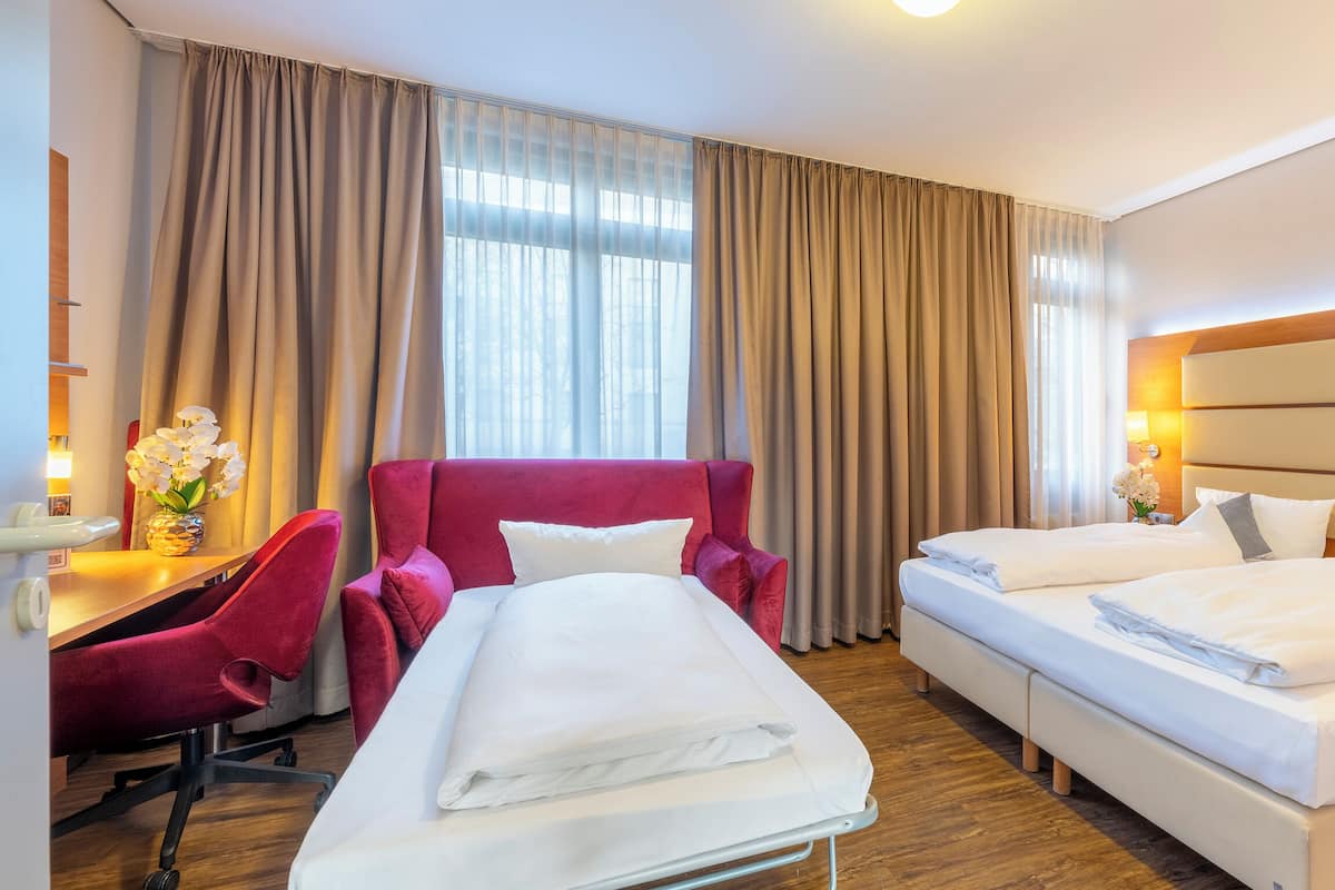 Habitación de hotel con cama, escritorio, silla y un ventanal con cortinas.