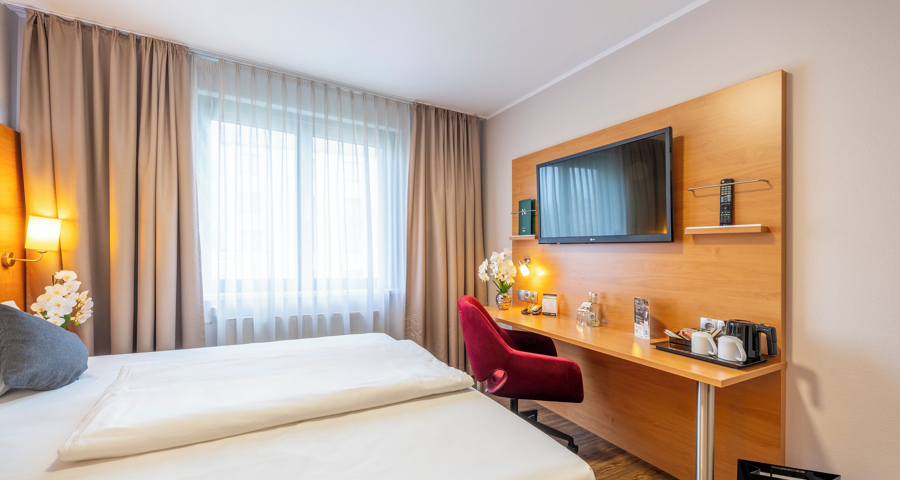 Habitación de hotel con dos camas, un escritorio con televisor, una silla roja y un jarrón con flores.