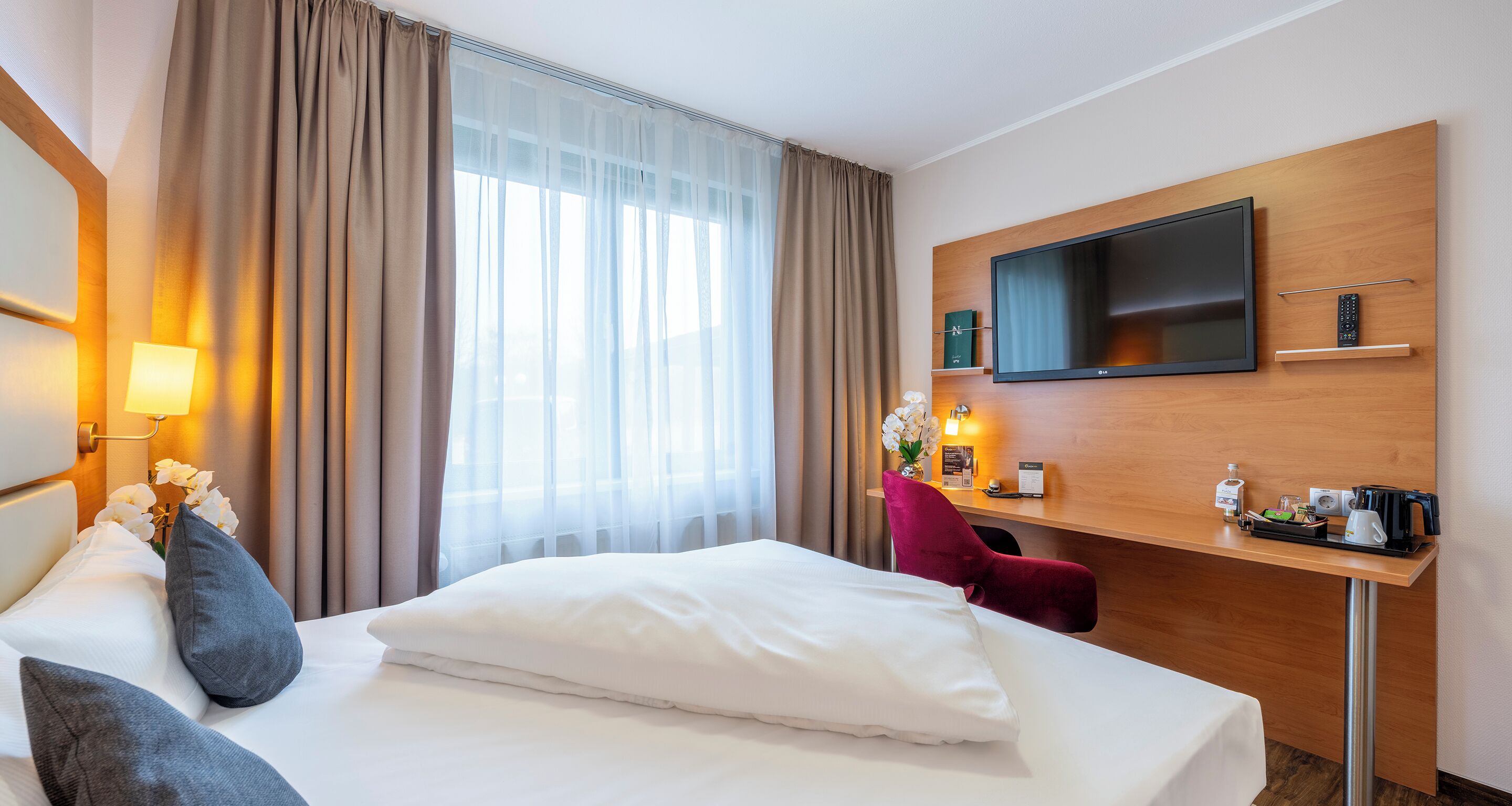 A hotel room with a bed, a desk with a TV, a chair, and a window with curtains.