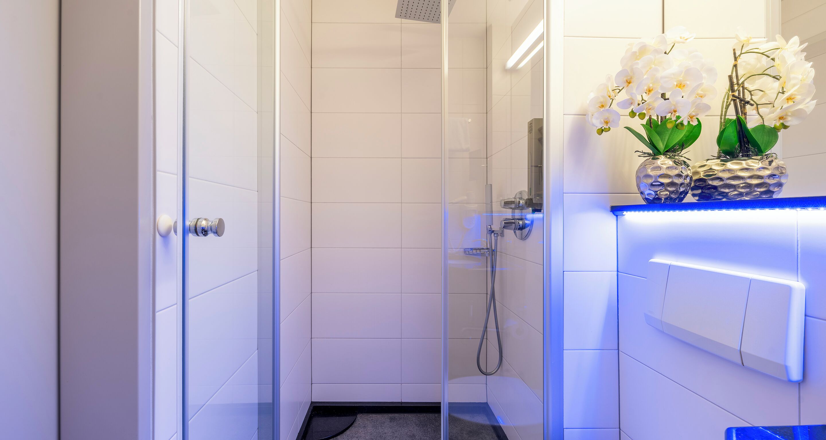 Une salle de bain moderne avec des carreaux blancs, une douche à l’italienne en verre, un pommeau de douche à main et un meuble-lavabo bleu éclairé par des diodes électroluminescentes, agrémenté d’orchidées.