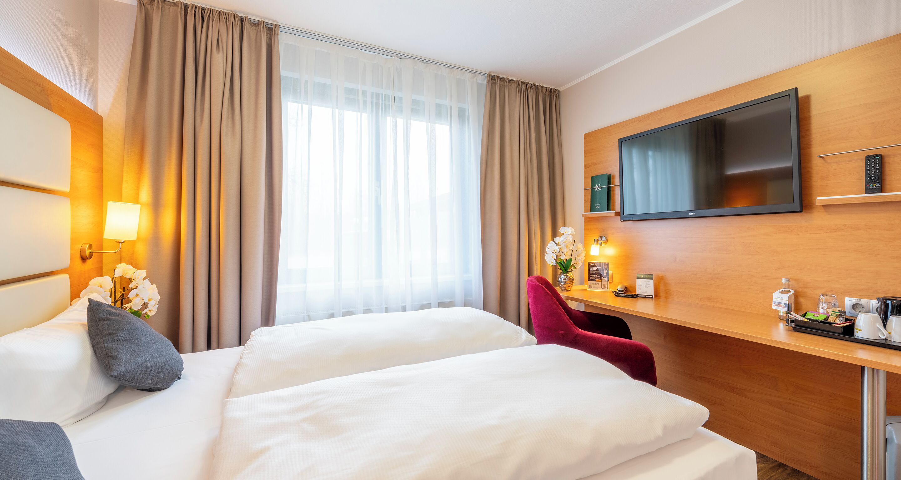 Une chambre d’hôtel avec deux lits, une télévision, un bureau avec une télécommande et une fenêtre avec des rideaux.