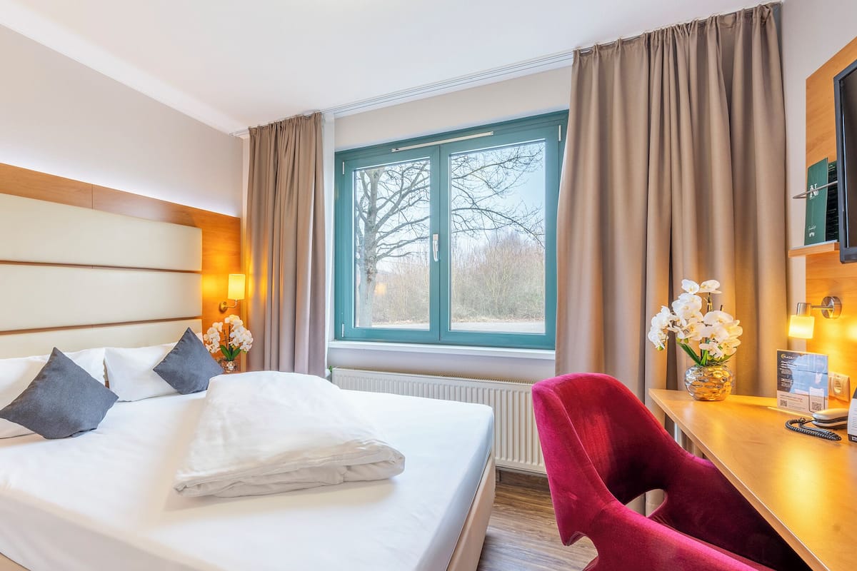 Una habitación de hotel con una cama grande, dos lámparas de noche, un escritorio con una silla y una ventana con cortinas.