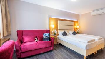 A hotel room with a red sofa, a bed with white linens, a nightstand with a lamp, and a wall-mounted air conditioner.