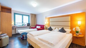 Une chambre d’hôtel avec deux lits, un bureau, une chaise, une fenêtre avec des stores et une tête de lit en bois.