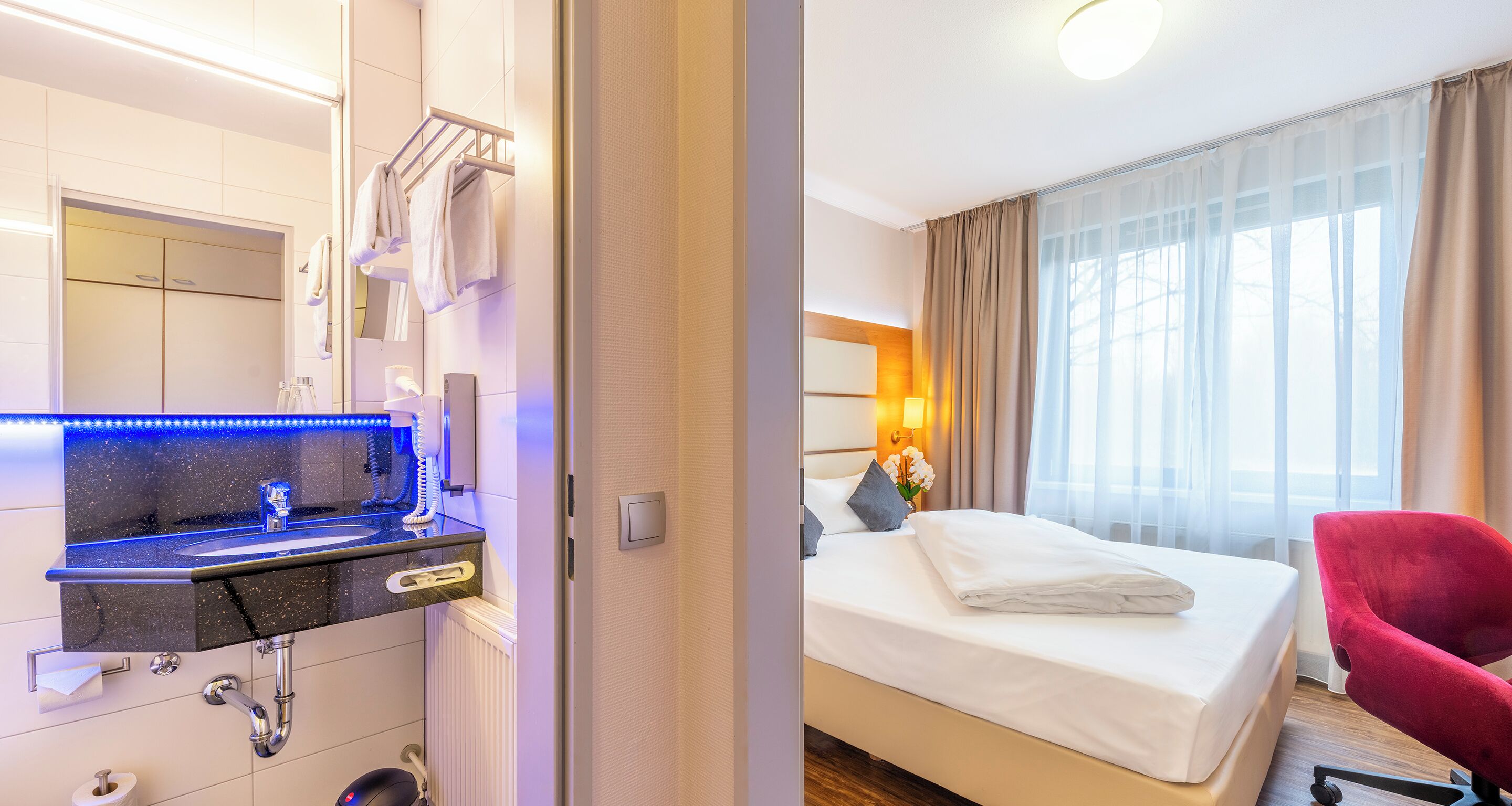 Ett hotellrum med en säng, en röd stol, ett badrum med handfat och spegel samt ett fönster med gardiner.