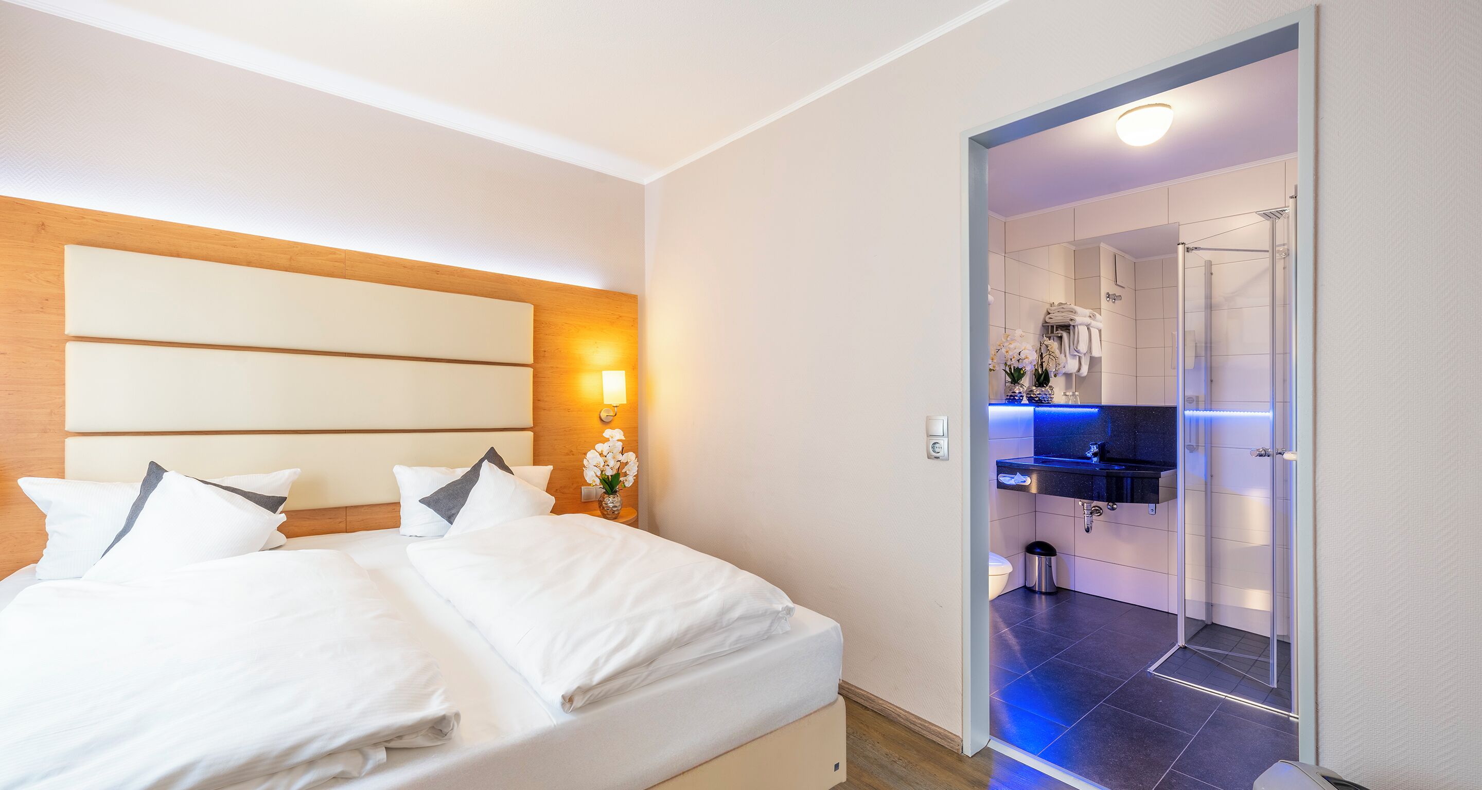 Une chambre d’hôtel avec un lit, une table de chevet, une lampe fixée au mur et une salle de bain visible à travers une porte ouverte.